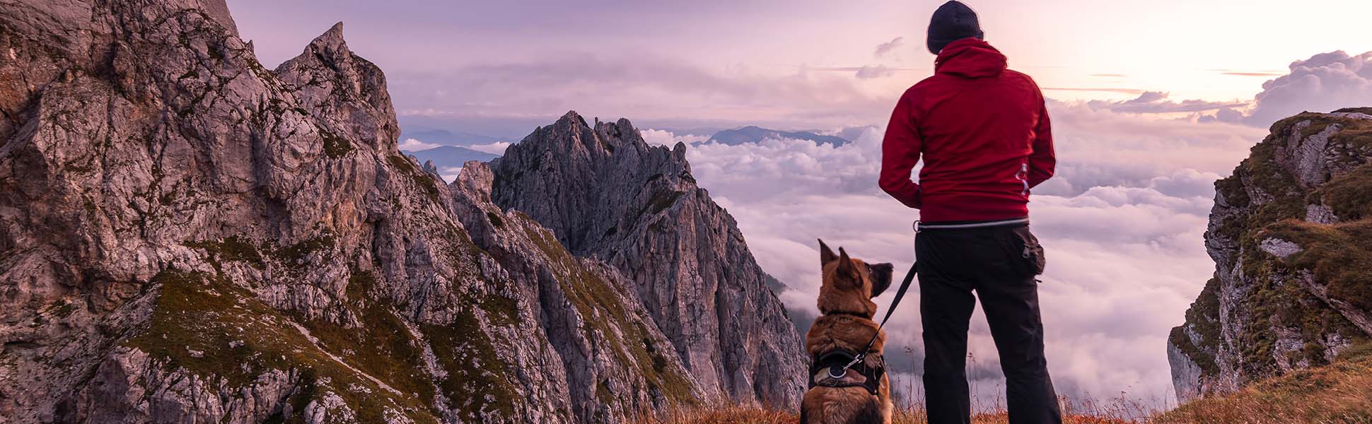 Man in red coat standing on a mountain trail with a german shepherd dog by his side. Man and dog are looking out at a beautiful sunrise over the clouds.